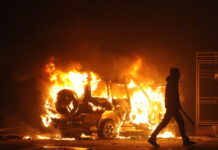 Burning vehicle at night, silhouetted person walking past.