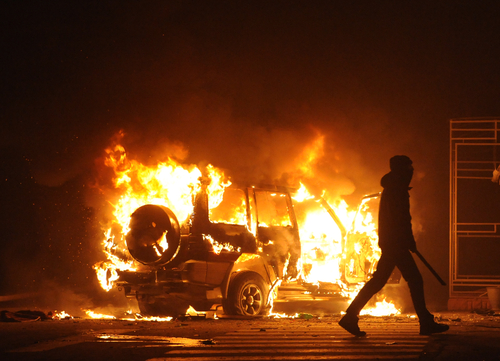 Burning vehicle at night, silhouetted person walking past.