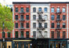 Red-brick building with fire escapes and storefronts, daytime.